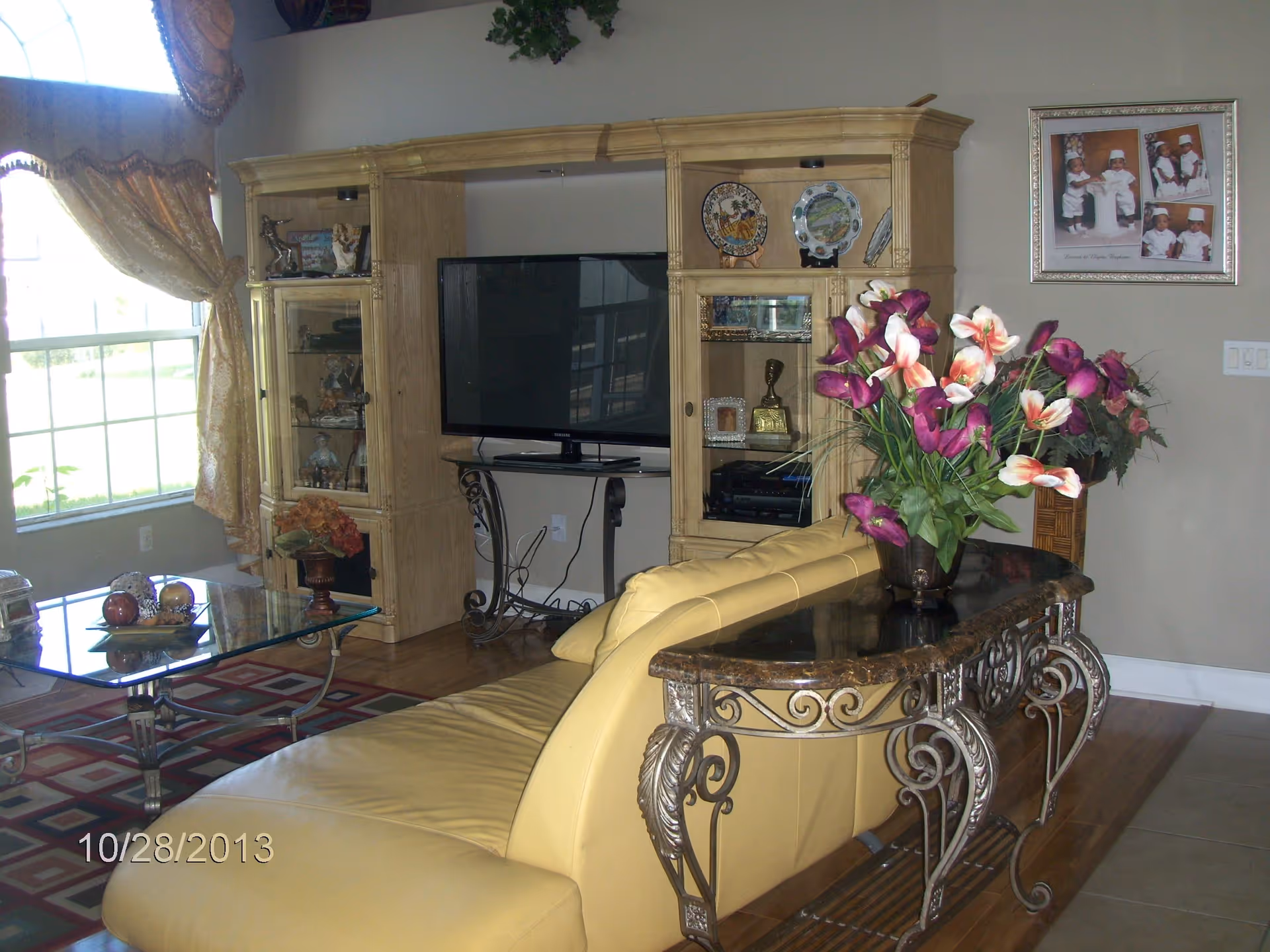 A cozy living room with a yellow leather couch, a decorative console table with a vase of purple and white flowers behind the couch, a glass coffee table with decorative items, a large window with gold curtains, and a wooden entertainment center holding a flat-screen TV and various decorative plates and figurines. A framed photo collage hangs on the wall.