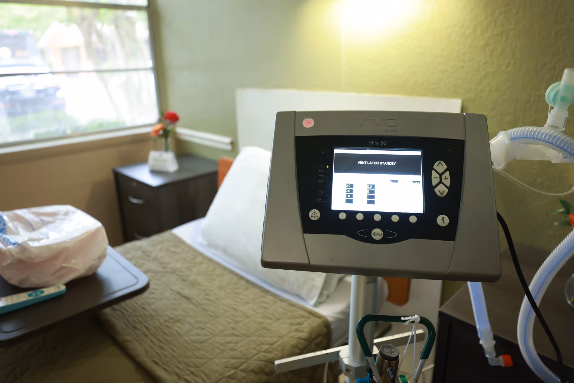 A medical ventilator machine in the foreground with a hospital bed in the background. The bed has a green blanket and white pillow, with a bedside table holding a small vase with red flowers. A window with a view of greenery is visible on the left side.