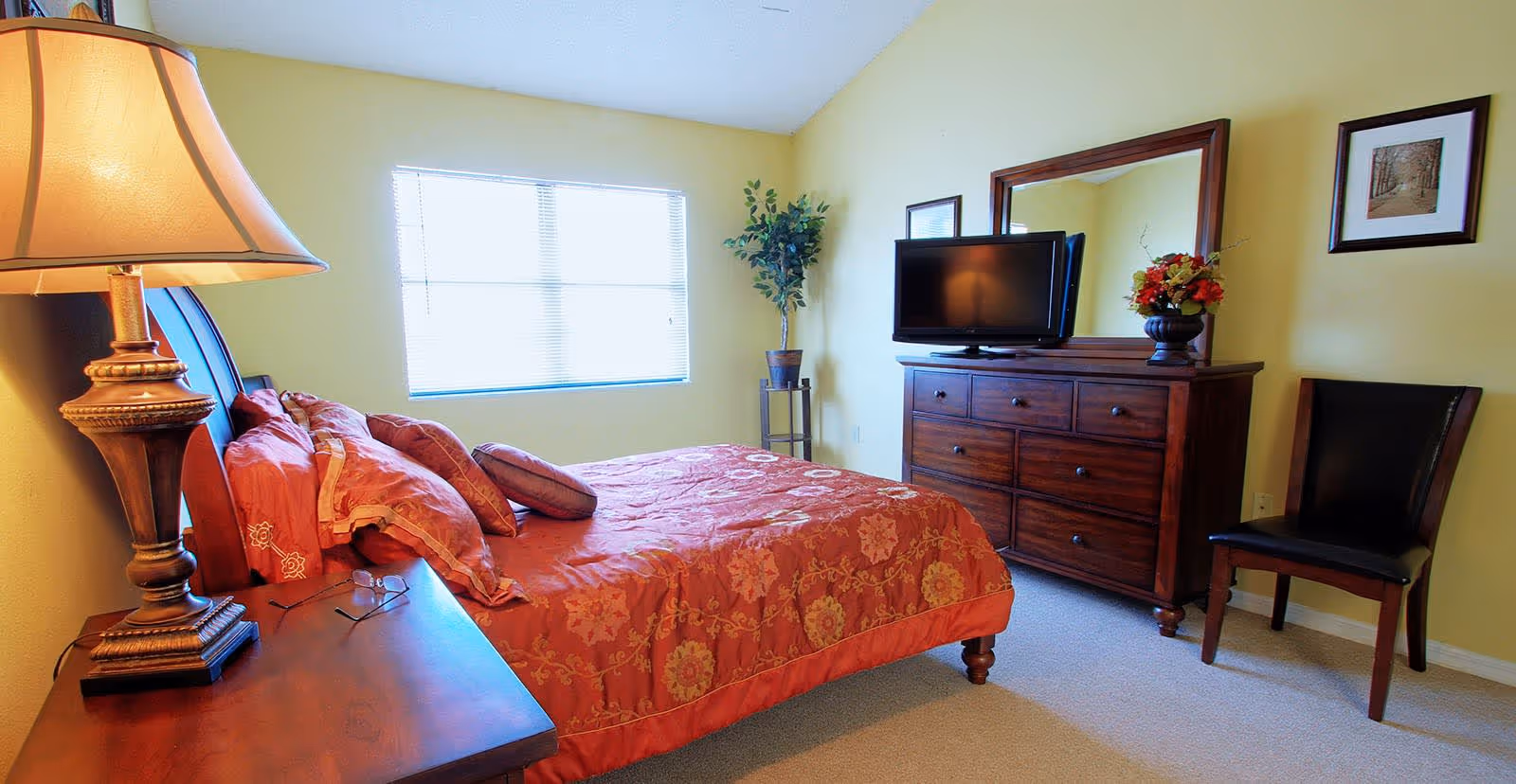 A bedroom with a large bed covered in a red and gold patterned comforter and multiple pillows. There is a wooden nightstand with a decorative lamp and a pair of glasses on it. Across from the bed is a wooden dresser with a flat-screen TV on top and a large mirror behind it. A black chair is placed next to the dresser, and a potted plant stands in the corner near a window with blinds. The walls are painted light yellow, and there are two framed pictures hanging on the wall.