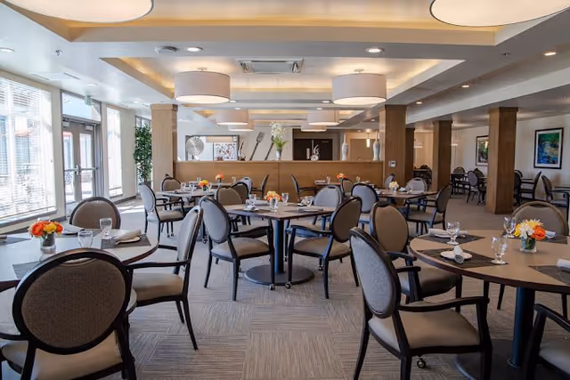 A spacious dining room with multiple round tables set with glasses, napkins, and small flower arrangements. The room features large windows allowing natural light, modern ceiling lights, and a neutral color palette with beige and brown tones.