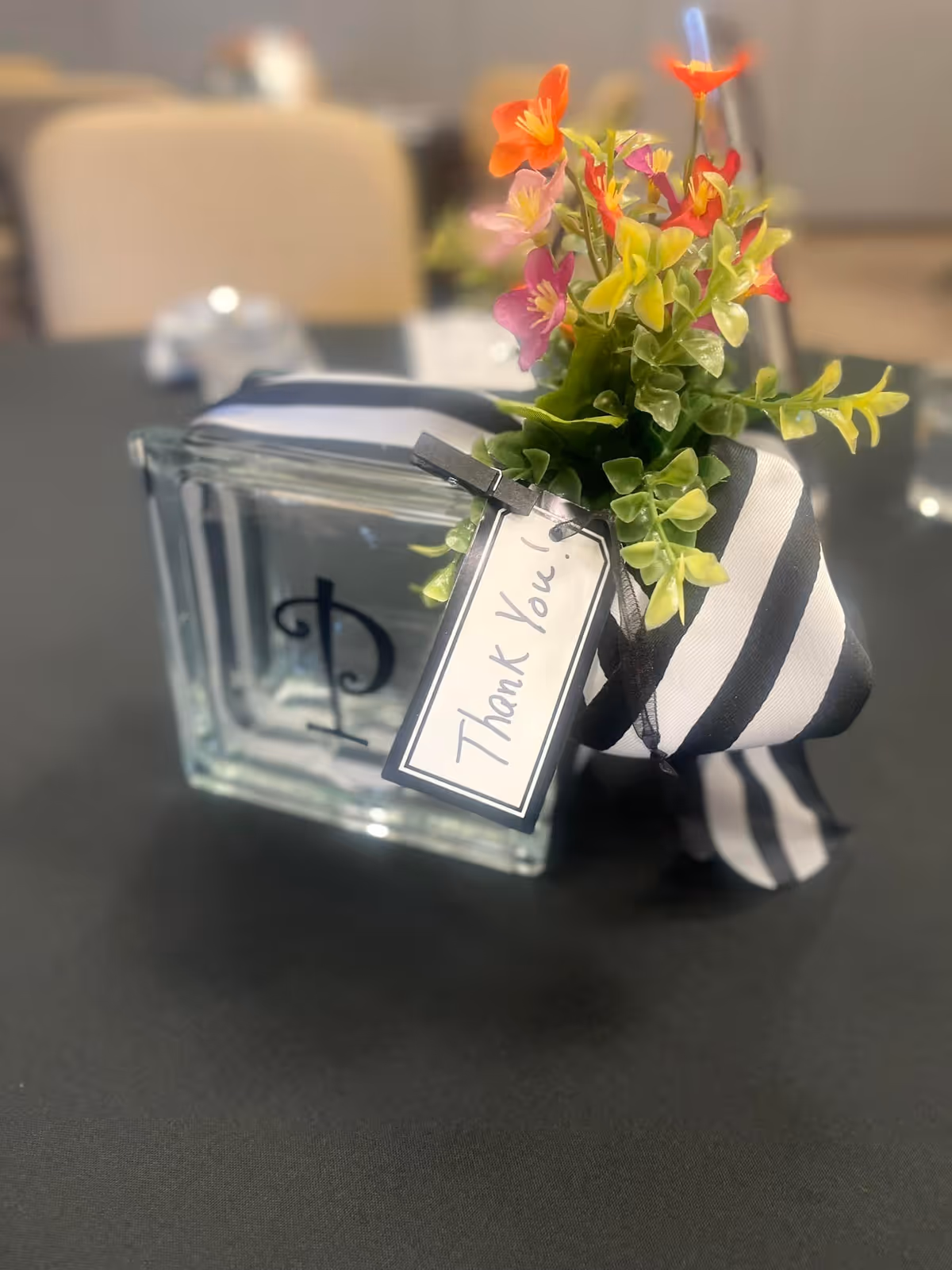 A glass block vase with a black letter 'P' on it, decorated with a black and white striped ribbon and a small bouquet of colorful artificial flowers. A tag attached to the ribbon reads 'Thank You!'. The vase is placed on a black tablecloth with blurred background elements.
