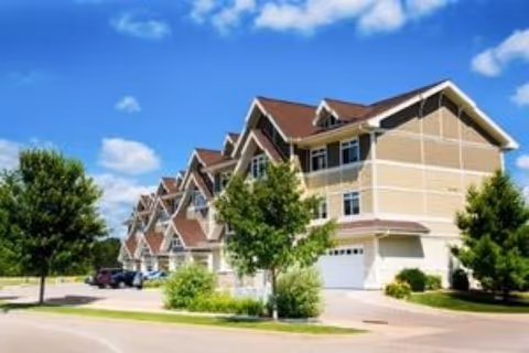 Multi-story residential building with pitched roofs, garages, trees and parked cars under a blue sky.