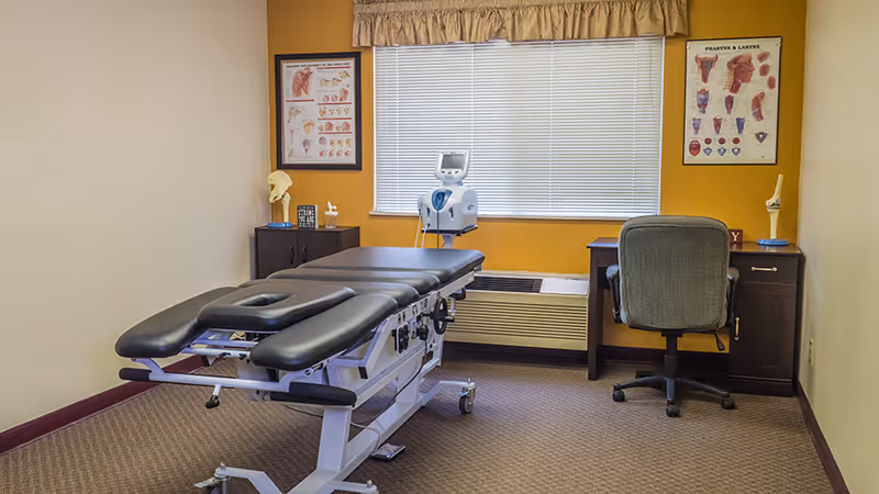 A medical examination room with a black padded examination table in the center, a desk with a chair facing away from the camera, medical posters on the walls, and a window with blinds partially closed. There is also a medical device on a stand near the window.