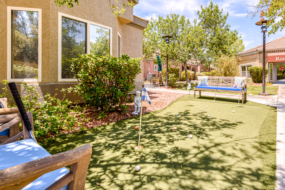 Outdoor putting green area at a senior living facility with benches, golf balls, flags, and surrounding greenery including bushes and trees under a sunny sky.