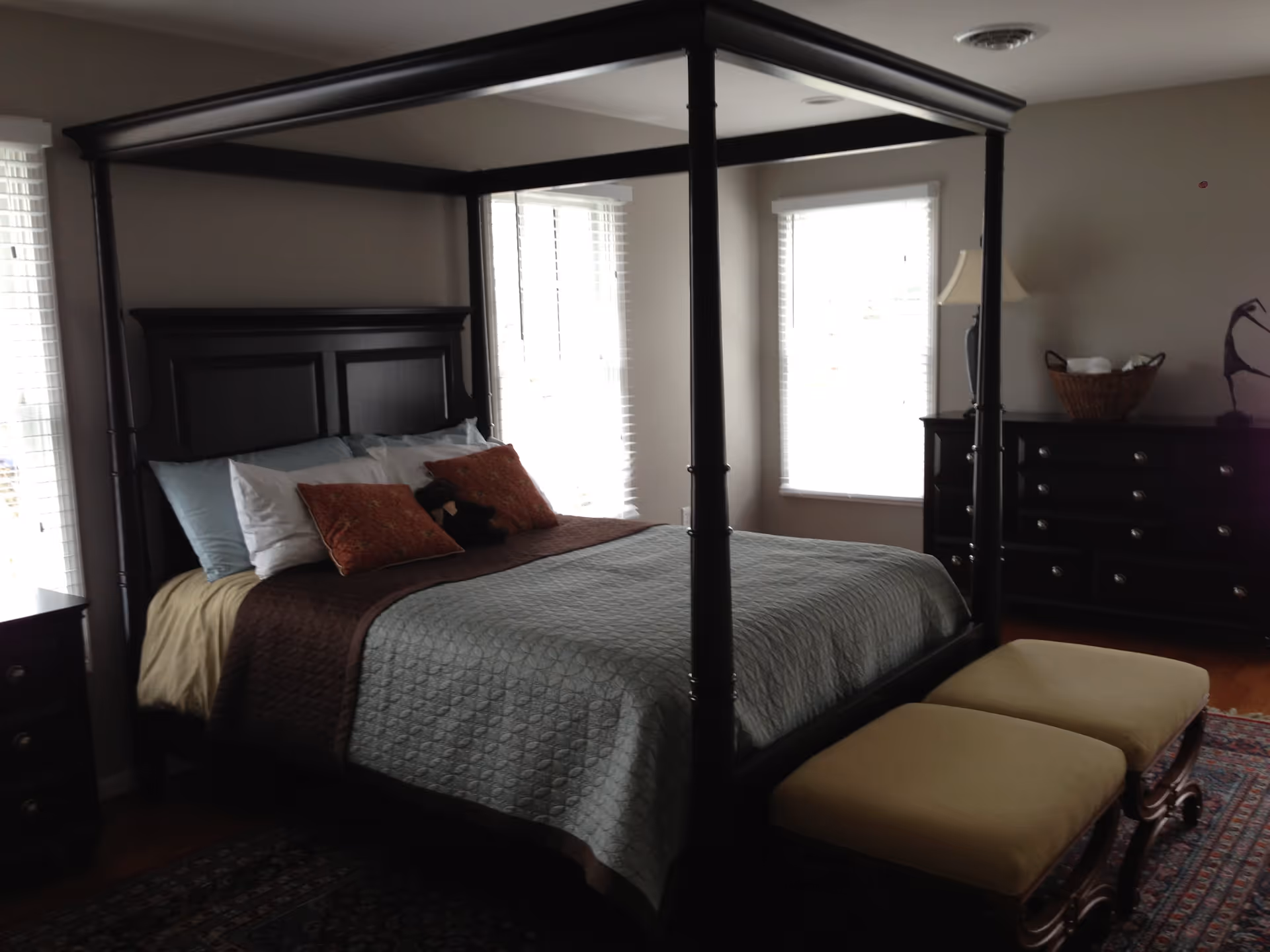A bedroom with a large dark wood four-poster bed made with light blue and brown bedding, several pillows, and a stuffed animal. The room has two windows with white blinds, a dark wood dresser with a lamp and a basket on top, and two beige cushioned benches at the foot of the bed.