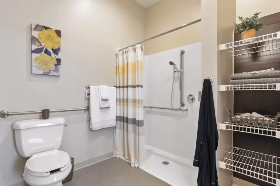 A clean and accessible bathroom with a white toilet, grab bars on the walls, a shower area with a handheld showerhead and a striped shower curtain. There are white towels hanging on a grab bar, wire shelving with folded towels and a potted plant, and a piece of wall art featuring yellow flowers.