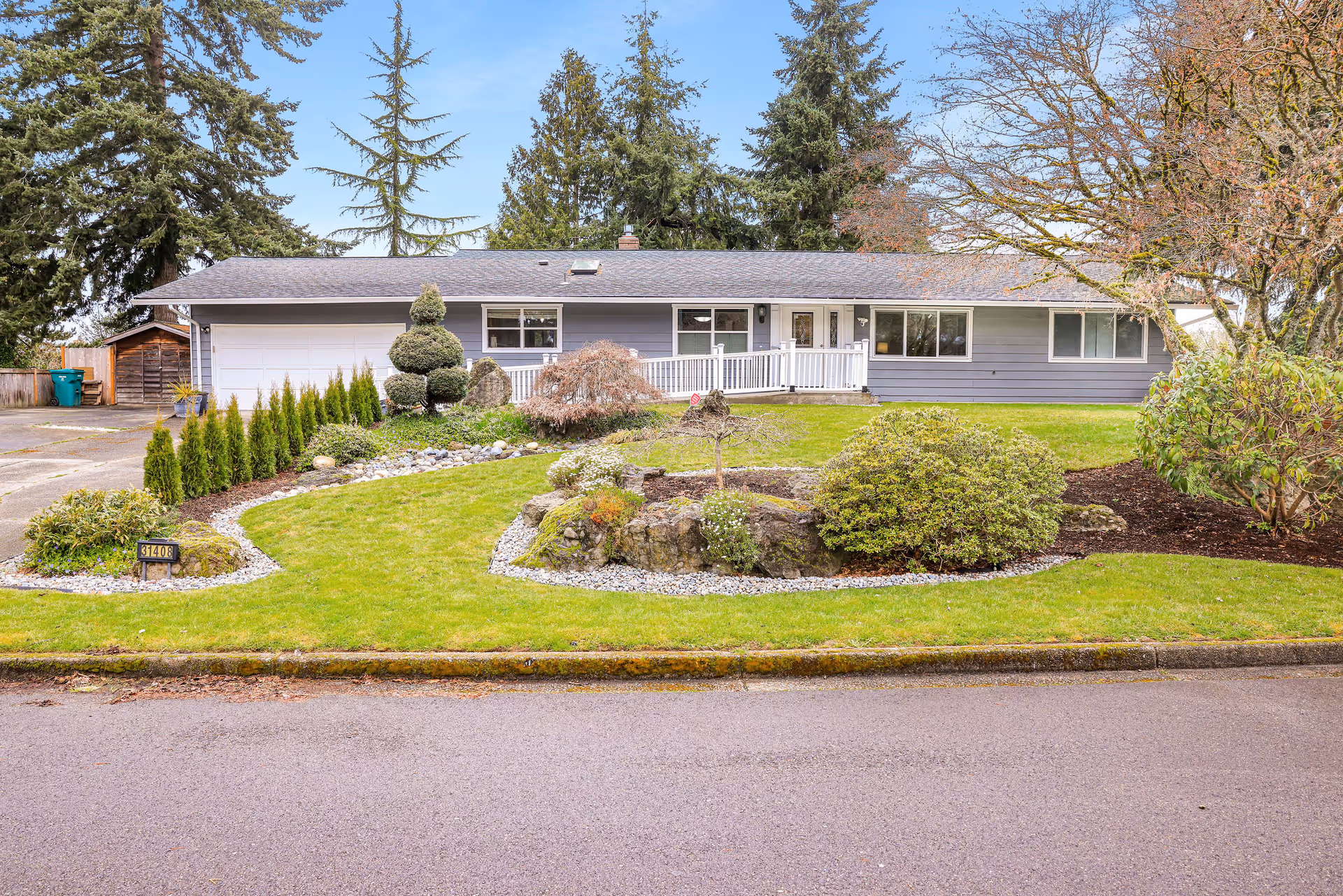 Single-story residential building with a gray exterior, white trim, and a front porch with a wheelchair ramp. The front yard is landscaped with green grass, various shrubs, small trees, and decorative rocks. Tall evergreen trees are visible behind the house under a clear blue sky.