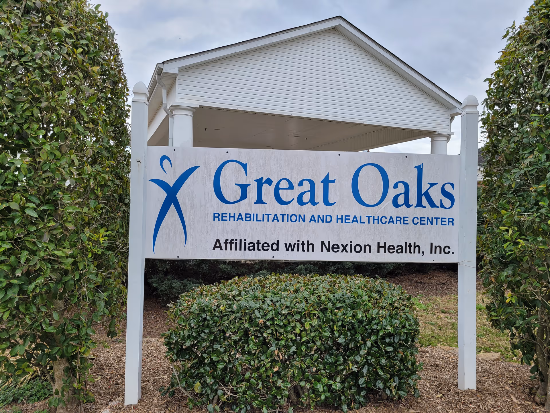 Outdoor sign for Great Oaks Rehabilitation and Healthcare Center, affiliated with Nexion Health, Inc., surrounded by green bushes and trees with a cloudy sky in the background.