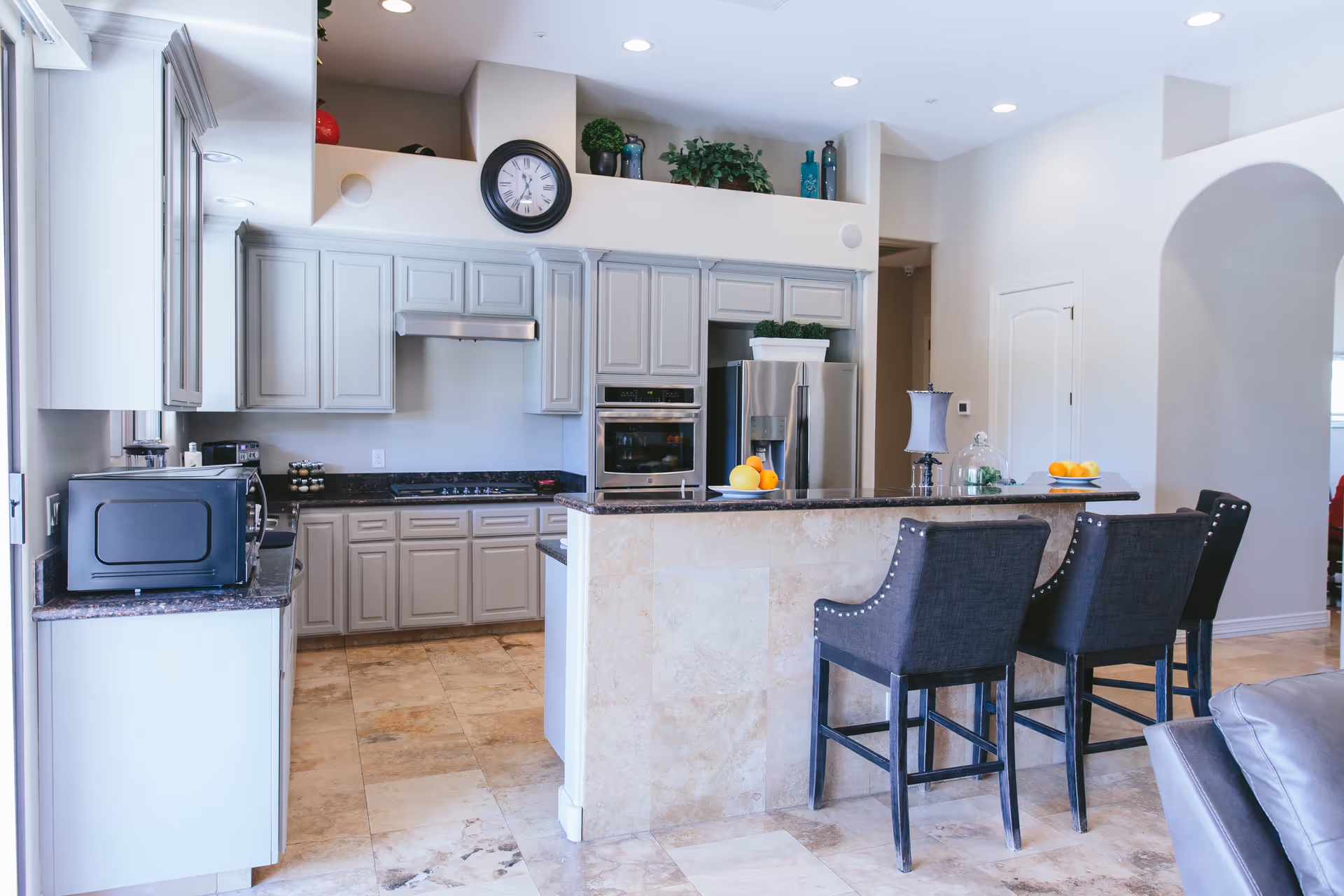A modern kitchen with light gray cabinets, a stainless steel refrigerator, built-in oven, and a stovetop with a range hood. There is a kitchen island with a dark countertop and three dark upholstered bar stools. Decorative plants and a clock are placed above the cabinets. The floor is tiled with beige stone tiles.