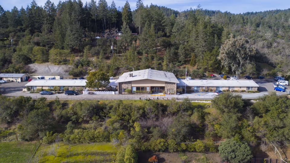 Aerial view of Cloverdale Healthcare Center, a single-story building with a light-colored roof surrounded by trees and greenery. Several cars are parked around the building, and the facility is set against a backdrop of forested hills under a clear sky.
