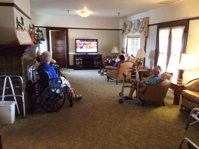 Several elderly residents in wheelchairs and armchairs sit in a communal living room watching a television.
