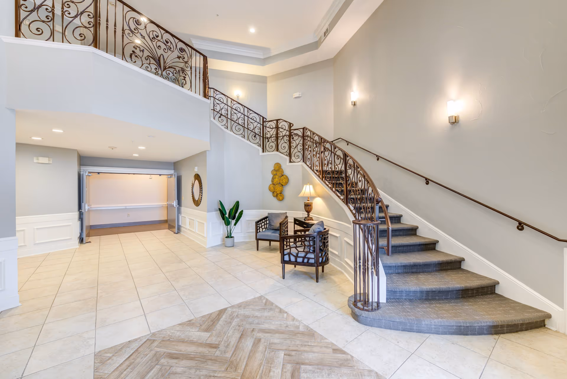 Interior view of a senior living facility lobby area featuring a curved staircase with ornate metal railings, two cushioned chairs with a small table and lamp between them, a potted plant, decorative wall art, and a large open doorway leading to another room. The floor is tiled with a combination of light tiles and wood-patterned tiles.