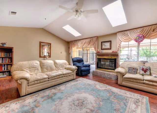 A cozy living room with two beige leather sofas and a blue recliner chair arranged around a fireplace. The room has a large floral area rug on wooden floors, a ceiling fan, skylights, and large windows with draped curtains letting in natural light. A bookshelf filled with books is visible in the corner.