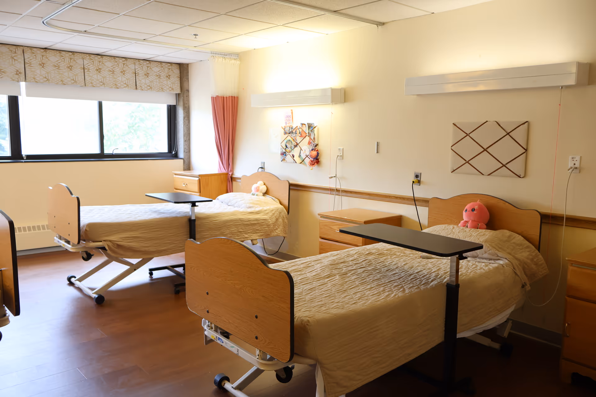 A bright room with two hospital-style beds, each with beige bedding and a small stuffed toy on the pillow. Each bed has an overbed table and a wooden nightstand beside it. The room has a large window with patterned curtains, light-colored walls, and wooden flooring.