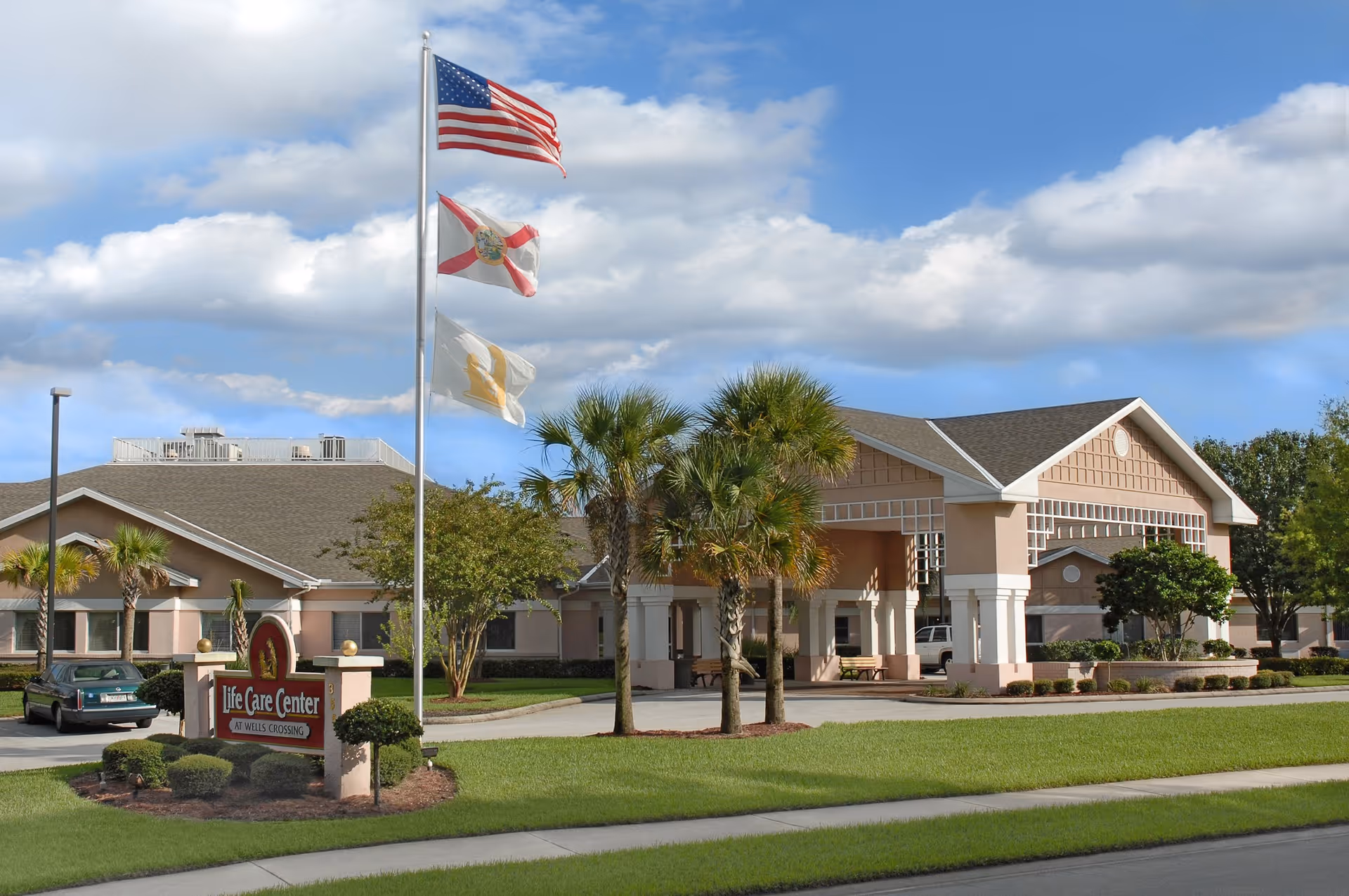 Exterior view of Life Care Center at Wells Crossing showing a single-story building with a covered entrance, palm trees, a green lawn, and three flags on a flagpole including the American flag. The sky is partly cloudy.