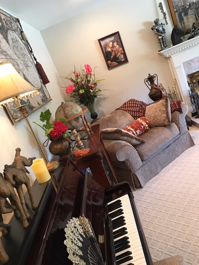 Cozy living room with a brown upholstered loveseat adorned with patterned pillows and a throw blanket. A piano with a decorative fan on the keyboard is in the foreground. The room features a floor lamp, a globe, a vase with flowers, and various decorative items including sculptures and framed artwork on the walls. A fireplace with a mantel and additional decorations is visible in the background.