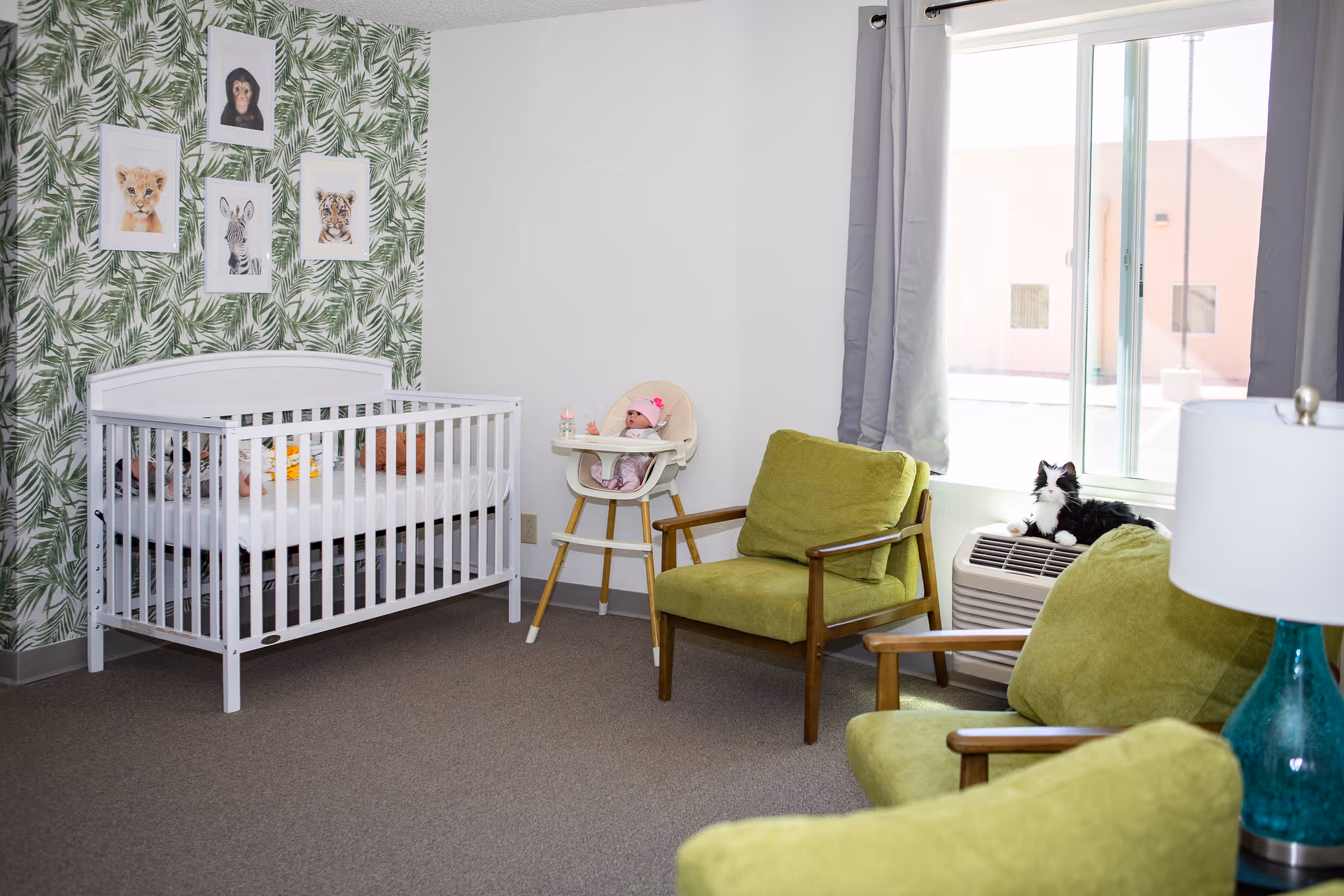 A cozy room with a white baby crib against a wall decorated with green leafy wallpaper and framed animal pictures. There is a wooden high chair with a doll seated in it, two green cushioned armchairs, a window with gray curtains, and a black and white cat sitting on an air conditioning unit below the window. A turquoise lamp is partially visible in the foreground.