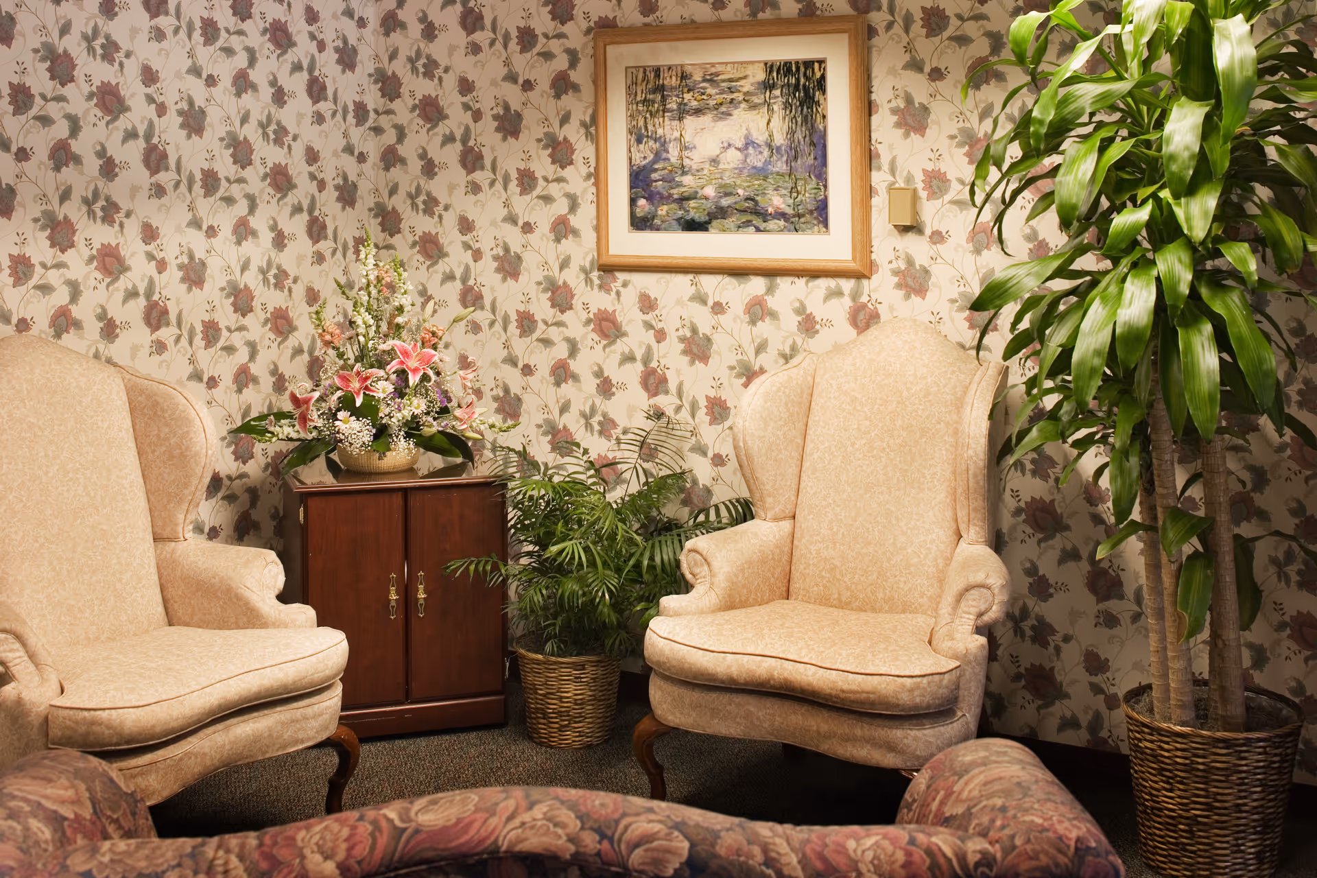 A cozy sitting area with two beige upholstered wingback chairs, a floral patterned sofa in the foreground, a wooden cabinet with a flower arrangement on top, two potted plants, floral wallpaper, and a framed painting on the wall.