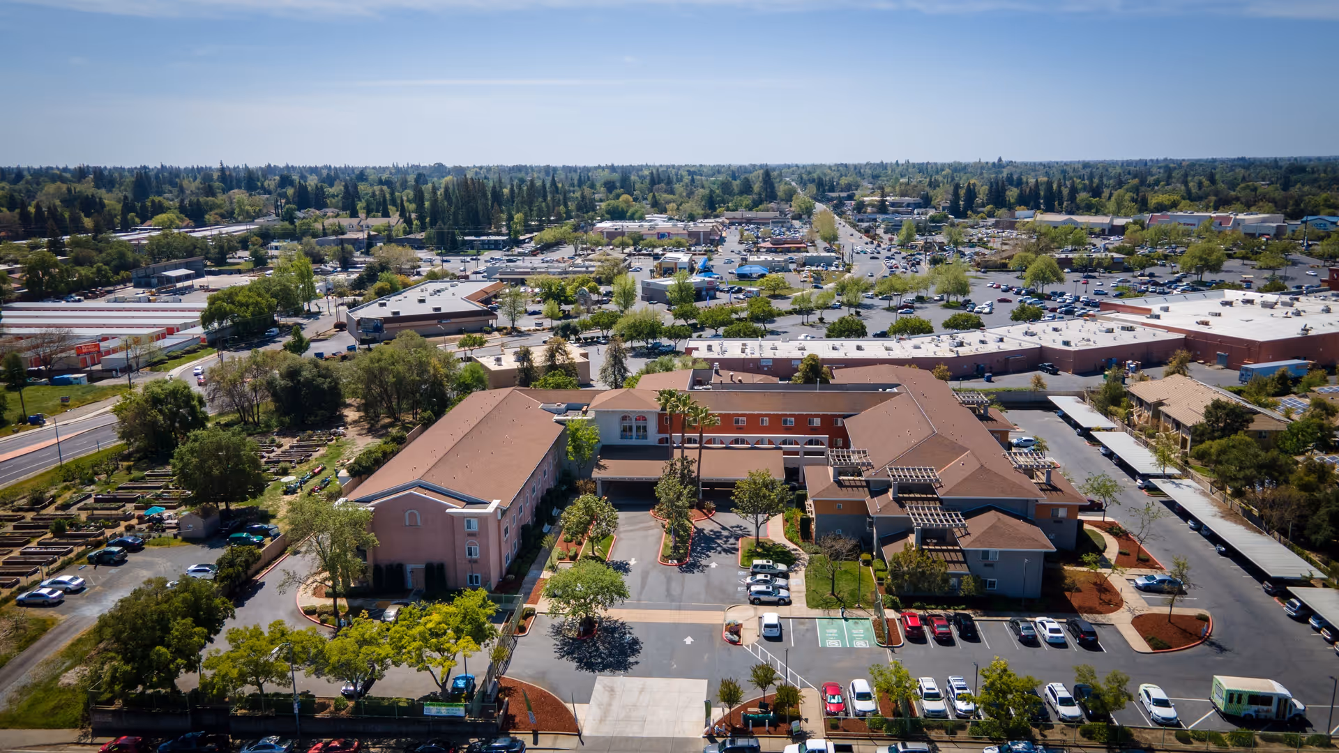 Aerial view of Cogir of Stock Ranch senior living facility showing multiple connected buildings with brown roofs, surrounded by parking lots, trees, and nearby commercial areas under a clear blue sky.
