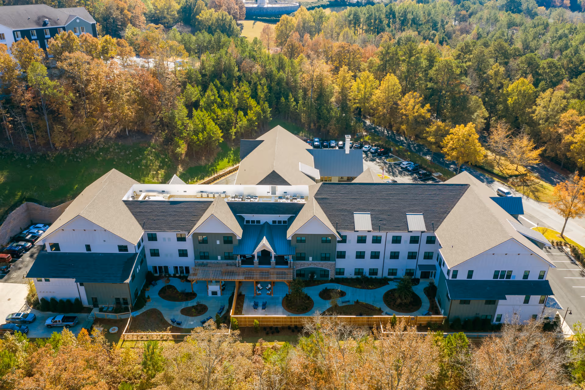 Aerial view of Longleaf Liberty Park senior living facility surrounded by trees with autumn foliage. The building is large with multiple sections and a courtyard area with pathways and landscaping. There are parking areas visible on the sides of the building.