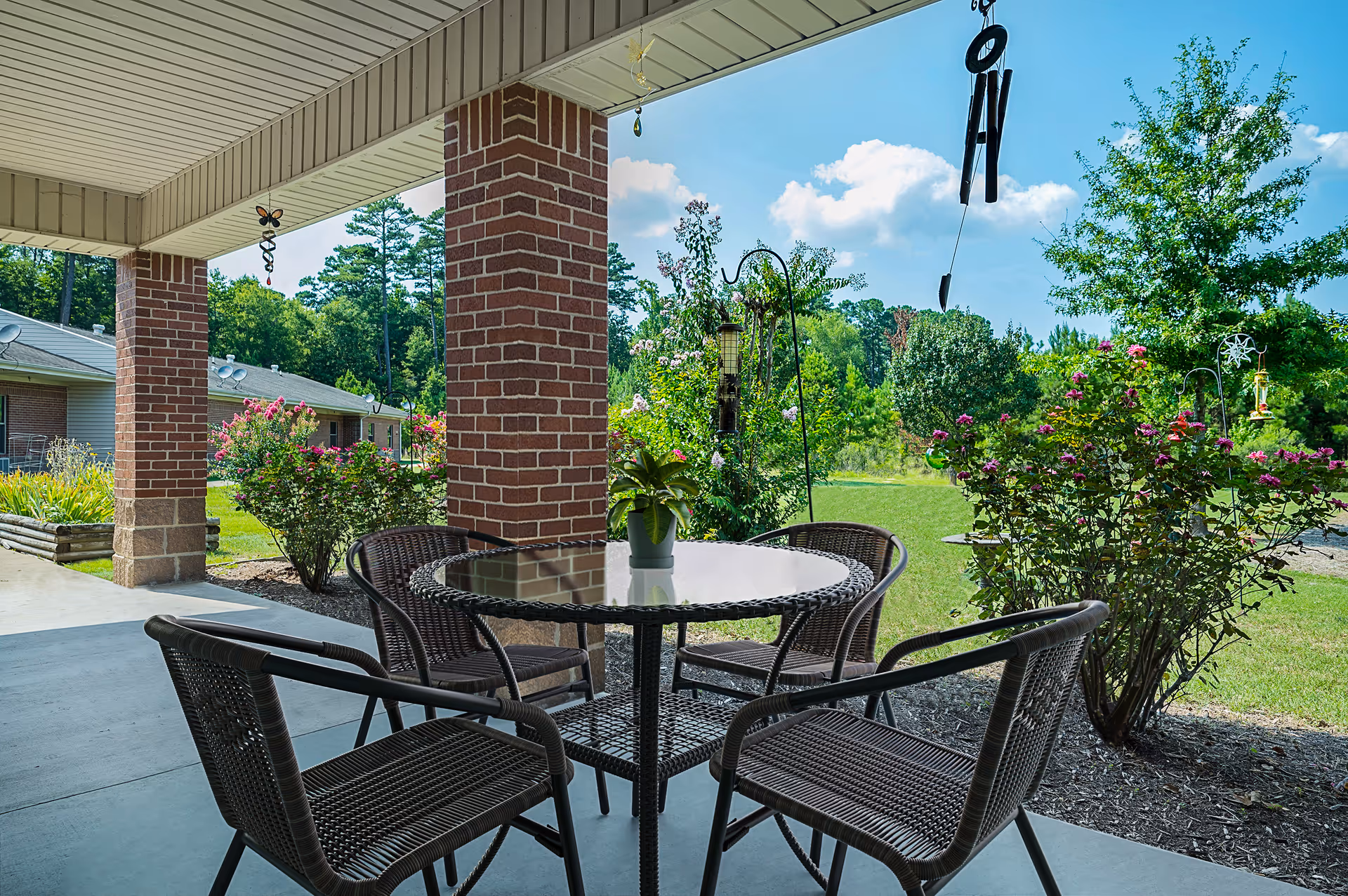 Covered patio with a round glass-top table and four wicker chairs overlooking a landscaped garden and lawn.