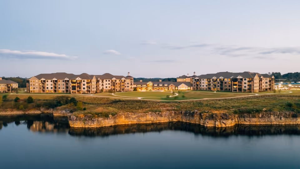 Row of senior living buildings on a grassy bluff overlooking a calm lake at dusk.