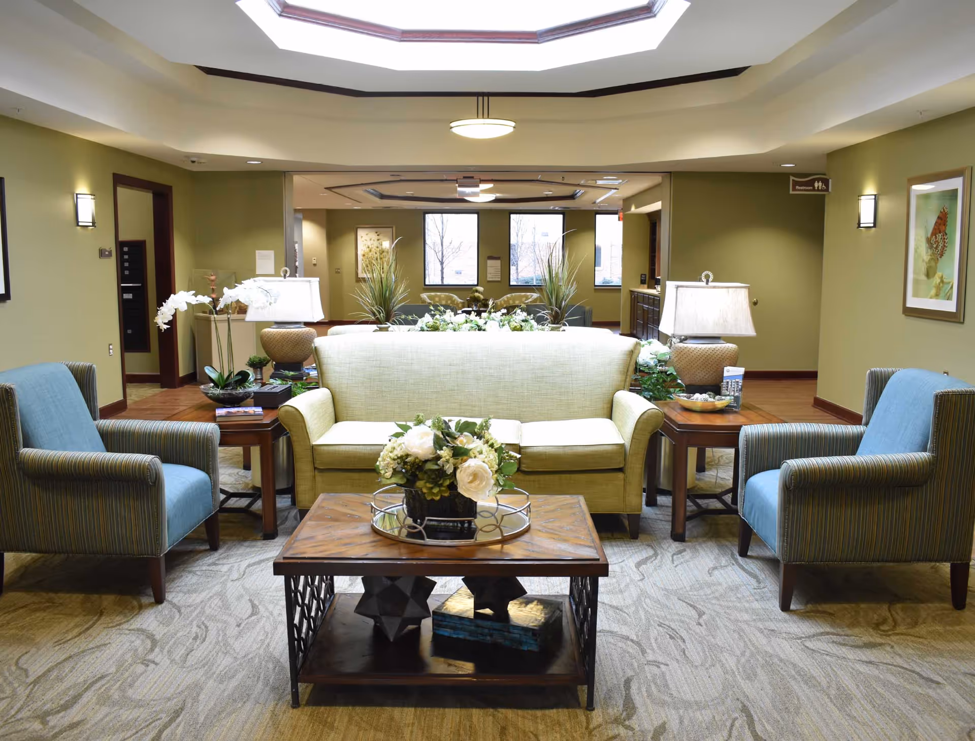 Bright seating lounge with a green sofa, two blue armchairs, a wooden coffee table with a flower arrangement, and side tables in a communal area.