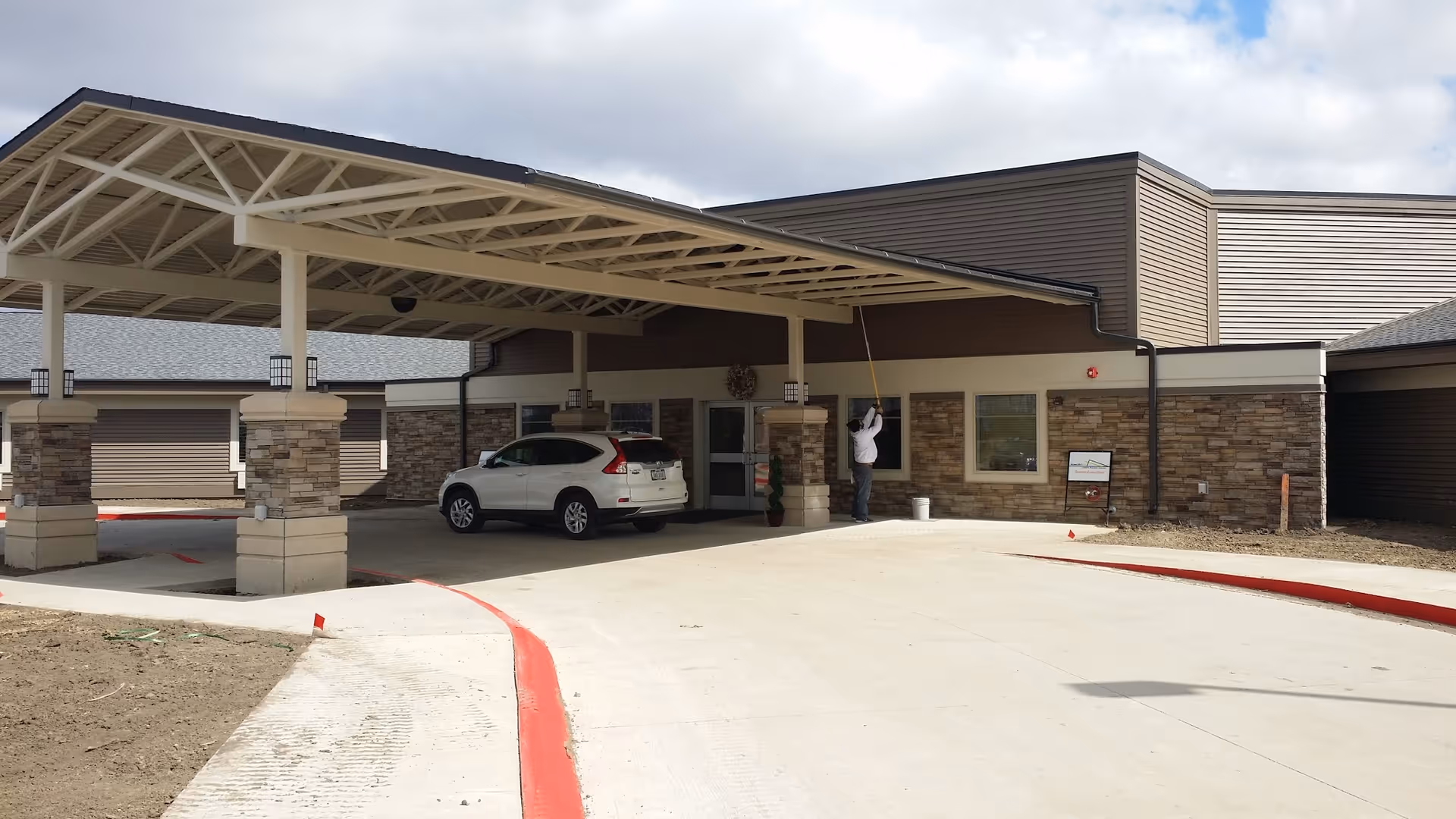 Entrance area of Pioneer Valley Living and Rehab facility with a covered drop-off zone supported by stone and beige pillars. A white SUV is parked under the canopy, and a person is cleaning or painting near the building's window. The building exterior features stone and brown siding with a concrete driveway and red-painted curbs.