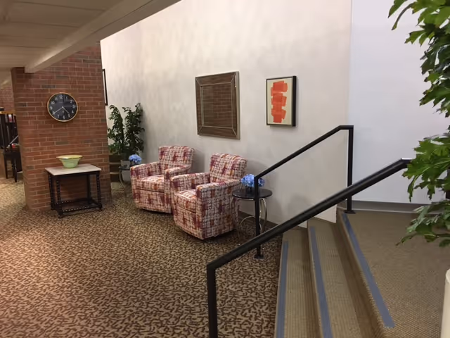 Lobby seating area with two patterned armchairs, side tables, plants, a clock on a brick pillar, and a short staircase with handrails.