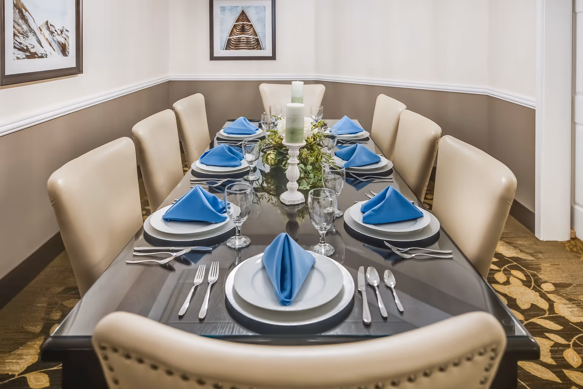 A formal dining room with a long dark wooden table set for eight people. Each place setting includes a white plate, silver cutlery, a water glass, and a blue folded napkin. The table centerpiece features white candles and green foliage. The room has beige walls with white trim and two framed pictures hanging on the walls.