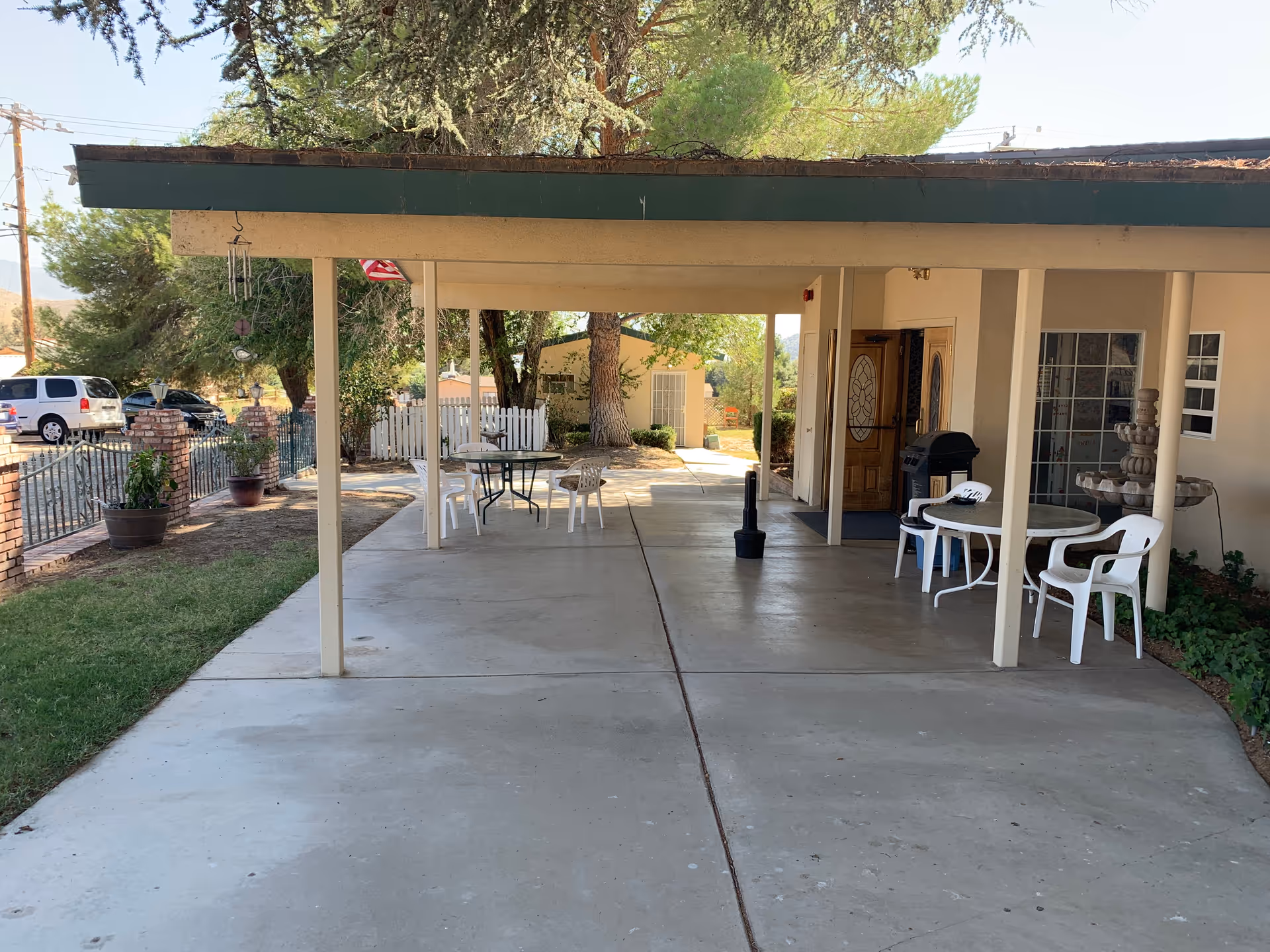 Covered outdoor patio area with white plastic chairs and round tables, a barbecue grill, and a decorative water fountain near the entrance of a building. Trees and a white picket fence are visible in the background along with parked cars and a brick fence.