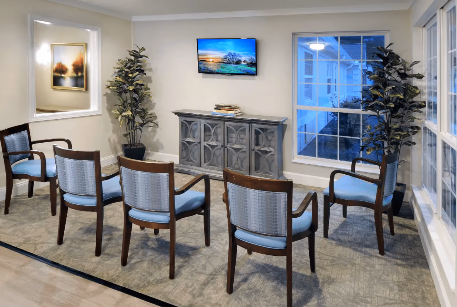 A small seating area with six wooden chairs featuring light blue cushions arranged in two rows facing a wall-mounted flat screen TV. Below the TV is a decorative gray cabinet with books on top. Two tall potted plants are placed on either side of the cabinet. Large windows on the right side let in natural light, and a large mirror hangs on the left wall reflecting a framed artwork.