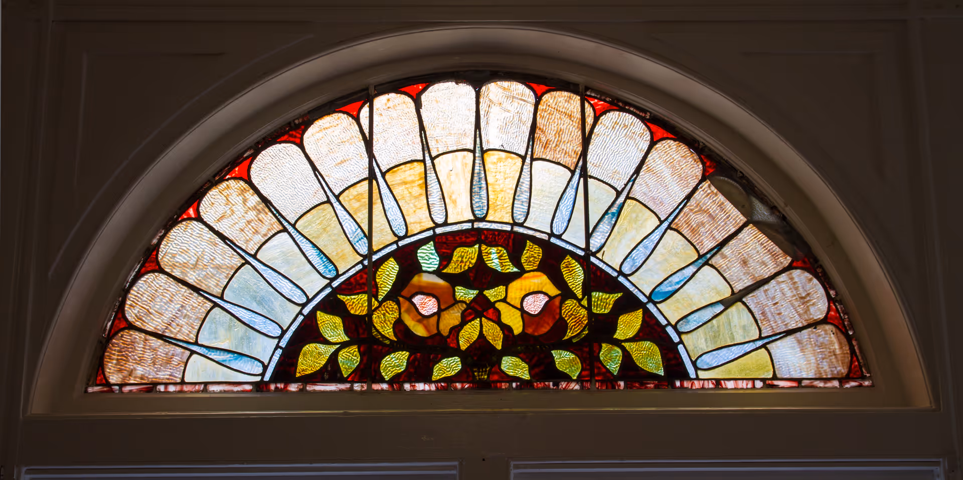 A semi-circular stained glass window featuring a floral and leaf design with various colors including red, yellow, green, and blue, set above a doorway.