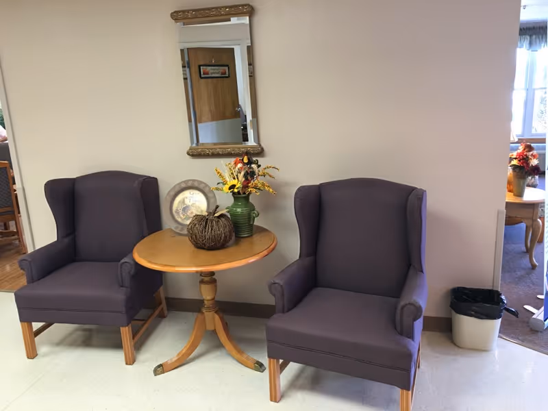 Two purple upholstered armchairs with wooden legs are positioned on either side of a small round wooden table. On the table, there is a decorative pumpkin, a green vase with yellow and orange flowers, and a decorative plate. A rectangular mirror with a gold frame hangs on the wall above the table. To the right, there is a small trash bin and a glimpse into another room with a window and additional furniture.