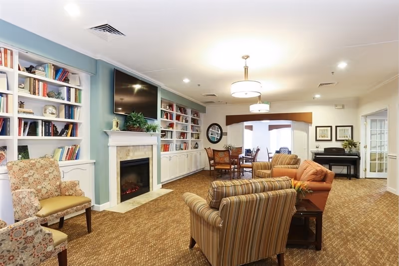 A cozy living room area in Oak Woods Manor featuring patterned armchairs and a sofa arranged around a fireplace with a mounted flat-screen TV above it. Built-in white bookshelves filled with books flank the fireplace. In the background, there is a dining table with chairs and a piano against the wall. The room is well-lit with ceiling lights and has a carpeted floor.