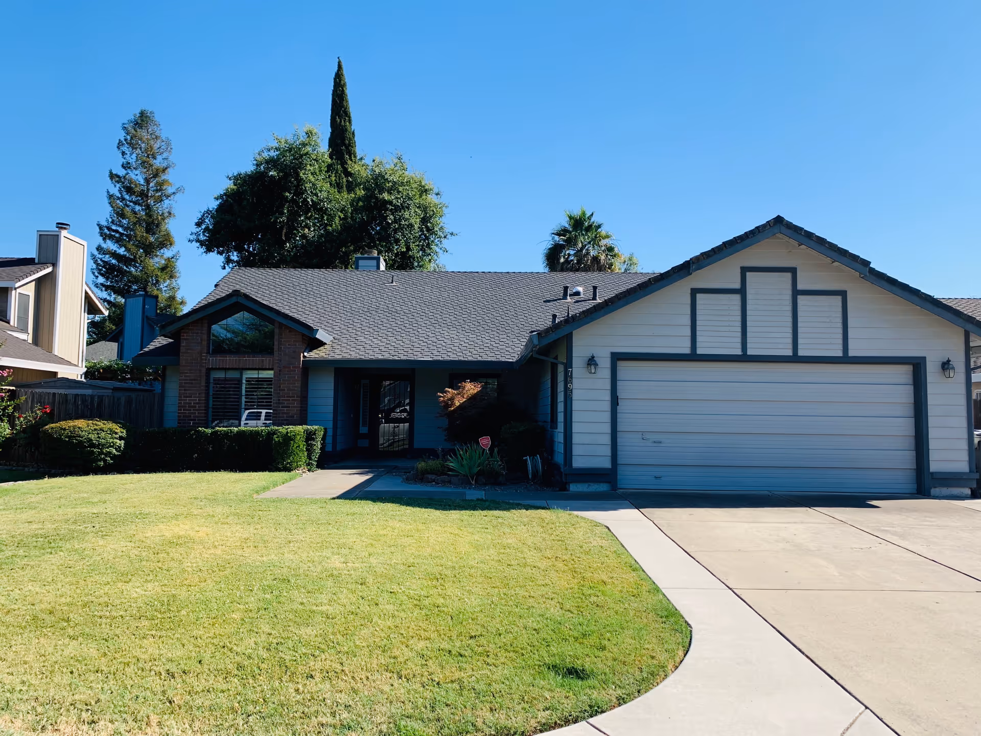 Single-story house with a two-car garage, a well-maintained front lawn, and various trees and shrubs under a clear blue sky.