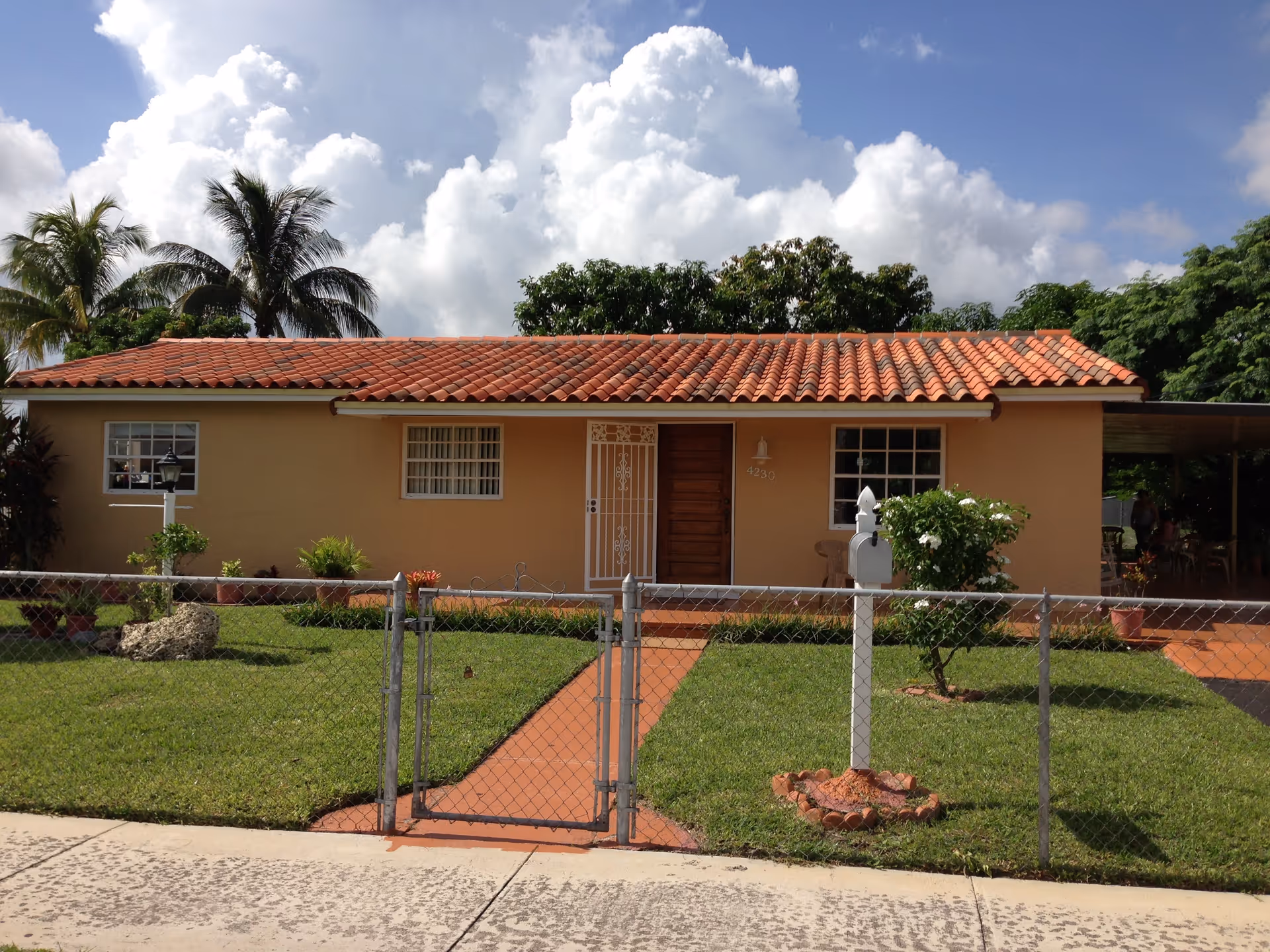 Single-story house with a red tiled roof, beige exterior walls, and a wooden front door with a white security door. The house has three windows with white frames and is surrounded by a well-maintained green lawn with a chain-link fence and a gate. There are palm trees and other greenery in the background under a partly cloudy blue sky.