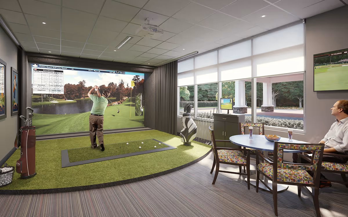 An indoor golf simulator room with a man swinging a golf club towards a large screen displaying a golf course. The room has artificial turf flooring in the golf area, a table with colorful chairs, and a man sitting and watching. Large windows with blinds let in natural light, and golf equipment is visible around the room.