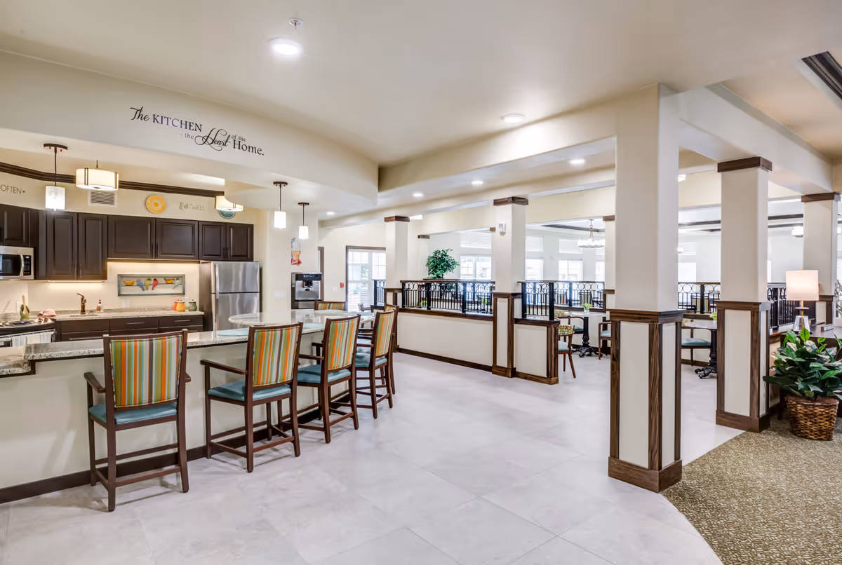 A bright and spacious kitchen and dining area in a retirement living facility. The kitchen features dark wood cabinets, a stainless steel refrigerator, microwave, and a granite countertop with four striped cushioned bar stools. Above the kitchen area, a decorative sign reads 'The Kitchen is the Heart of the Home.' The dining area has multiple tables and chairs with large windows allowing natural light to fill the space. The room has light-colored tiled floors and cream-colored walls with wooden trim accents.