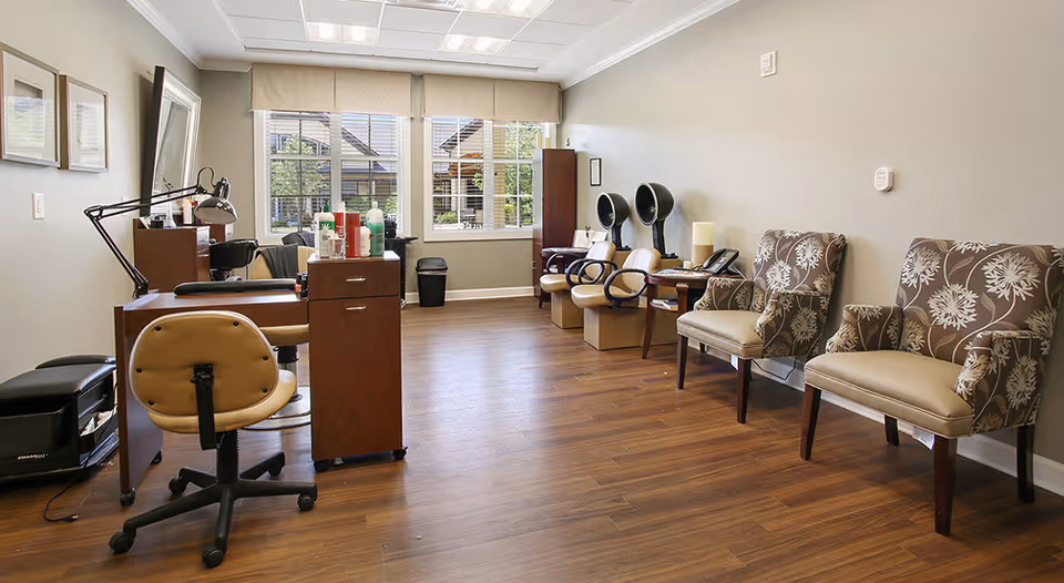Interior salon area with manicure stations, hooded hair dryers, and patterned armchairs facing large windows.