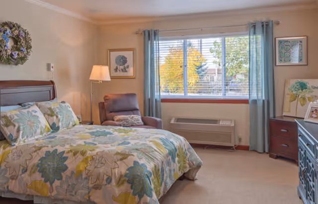 A cozy bedroom with a bed featuring floral patterned bedding in shades of green, blue, and yellow. There is a comfortable armchair with a pillow next to a floor lamp. The room has a large window with blue curtains, letting in natural light and showing trees outside. The walls are decorated with framed artwork and a wreath above the bed. A wooden dresser and a cabinet are also visible.