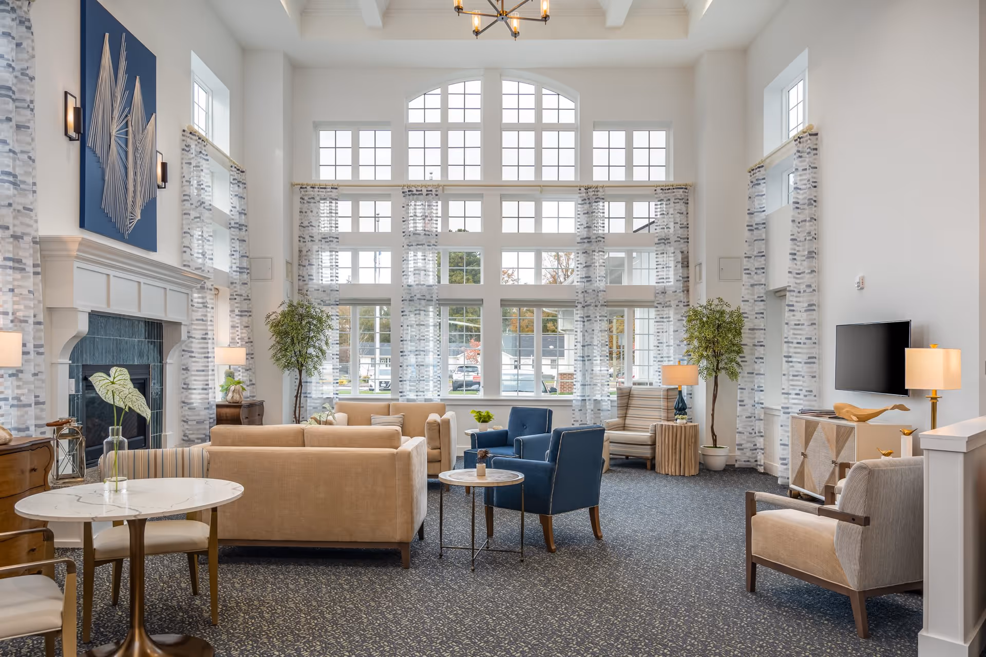 A bright and spacious senior living community lounge with large windows featuring white frames and sheer patterned curtains. The room has high ceilings with exposed beams and a modern chandelier. There are beige sofas, blue armchairs, and a striped armchair arranged around small tables. A fireplace with a blue abstract wall art above it is on the left side, and a flat-screen TV is mounted on the right wall. Several lamps and potted plants add warmth and greenery to the space.