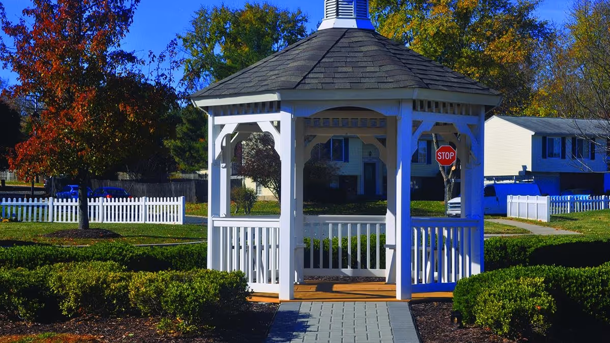 A white wooden gazebo with a shingled roof situated in a landscaped outdoor area with green bushes and trees showing autumn colors. In the background, there are residential buildings, a white picket fence, a stop sign, and parked vehicles under a clear blue sky.