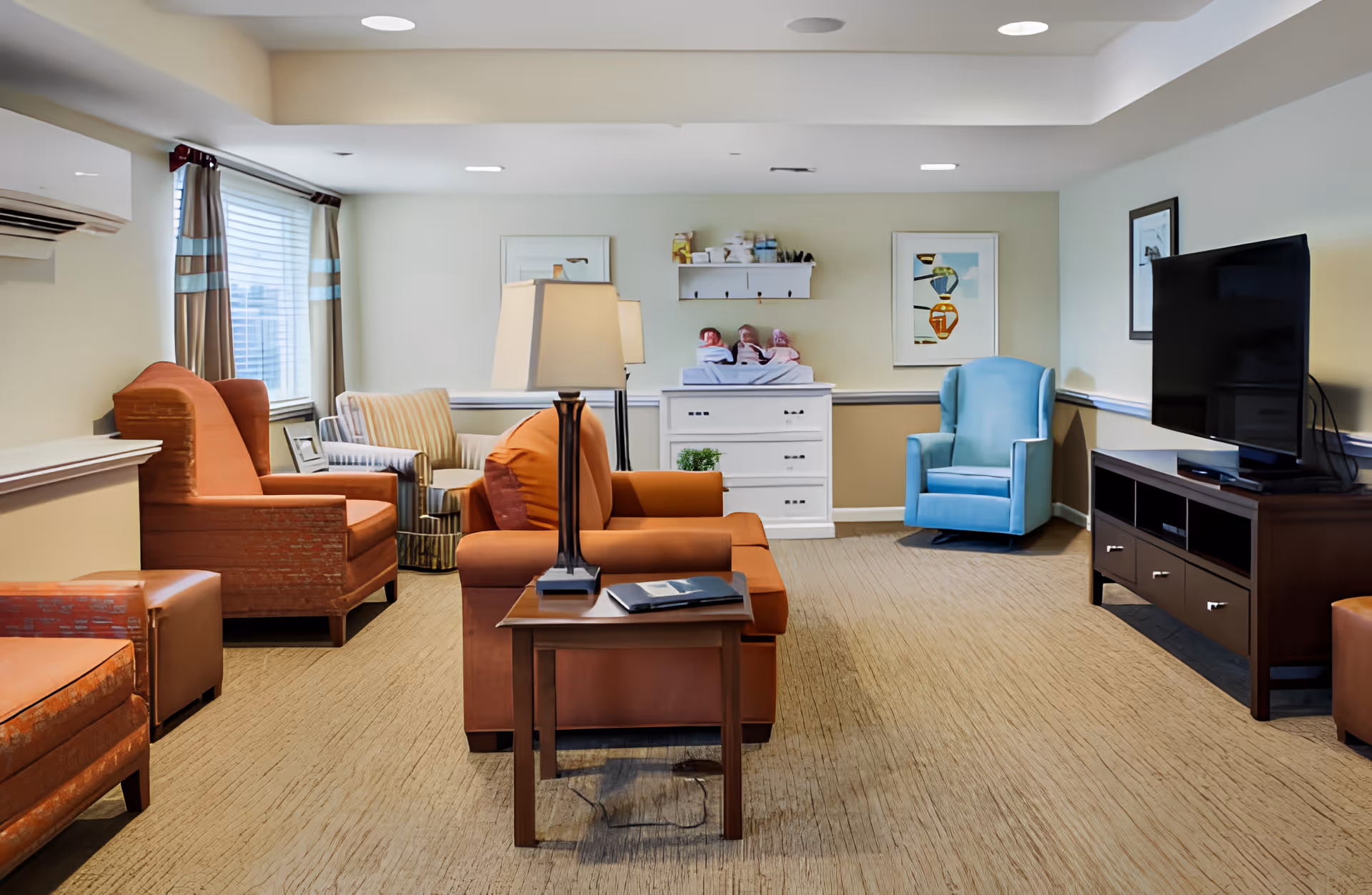 A bright memory-care living room with upholstered armchairs and sofas, a TV on a console, side tables and a dresser against the back wall.