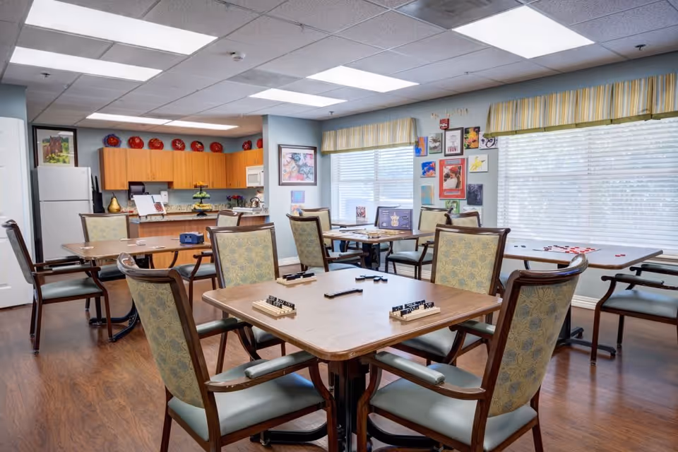 A bright and spacious common area with several wooden tables and cushioned chairs arranged for playing games. The tables have game pieces and racks on them. The room has large windows with striped valances, colorful artwork on the walls, and a kitchen area with wooden cabinets and a white refrigerator in the background.