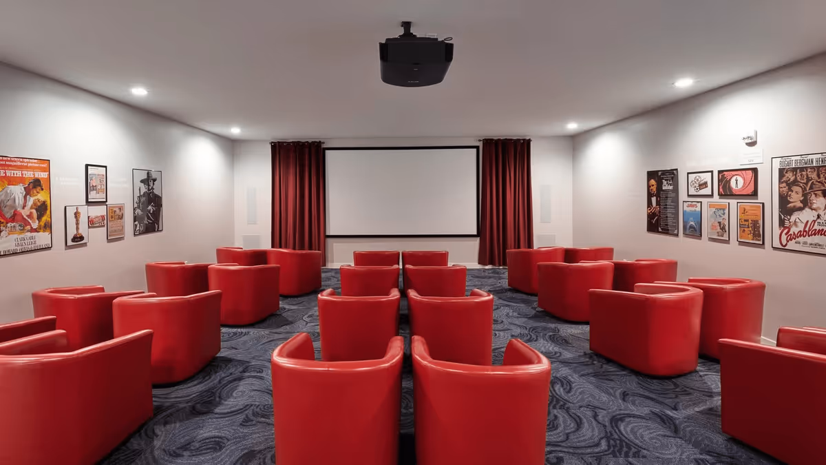 A small movie theater room with red leather armchairs arranged in rows facing a large white projection screen. The walls are decorated with framed classic movie posters, and the room has a dark patterned carpet and recessed ceiling lights.