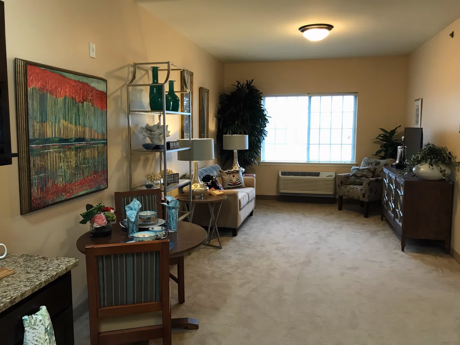 A cozy living room area with beige walls and carpet. There is a small round dining table with two chairs set with bowls, plates, and napkins. A large colorful abstract painting hangs on the left wall. A metal shelving unit holds decorative items including green vases and coral. A beige sofa with pillows and a side table with a lamp are positioned near a large window with blinds. Across from the sofa is a patterned armchair and a wooden cabinet with a TV on top. Several plants add greenery to the room.