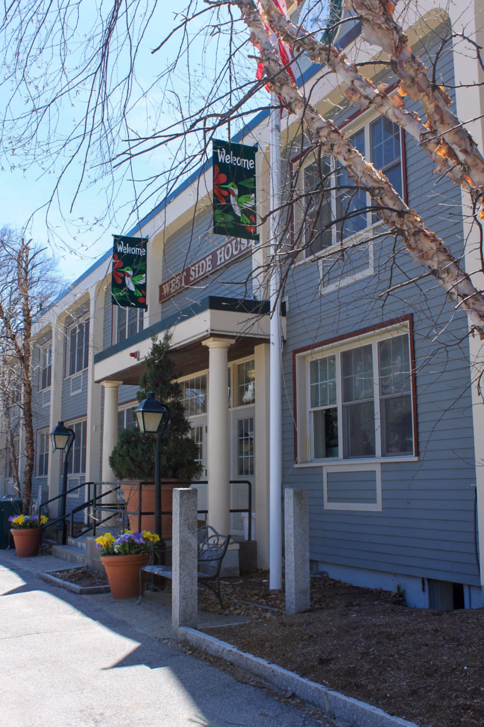 Exterior view of West Side House of Worcester, a two-story building with blue siding and white trim. The entrance features white columns, a small porch, and two welcome banners hanging above. There are potted plants with flowers and a bench near the entrance. Leafless trees are visible in the foreground and background.