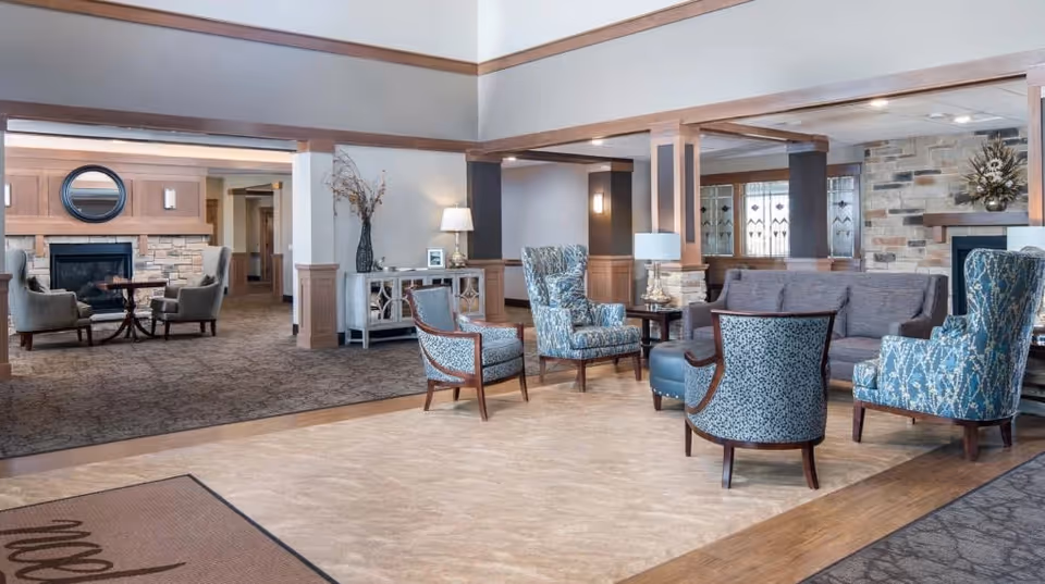 A spacious and well-lit living room area in a retirement facility featuring comfortable upholstered chairs and sofas arranged around a central ottoman. The room has wooden trim accents, a stone fireplace, decorative vases, lamps, and a carpeted floor transitioning to a tiled area near the entrance.