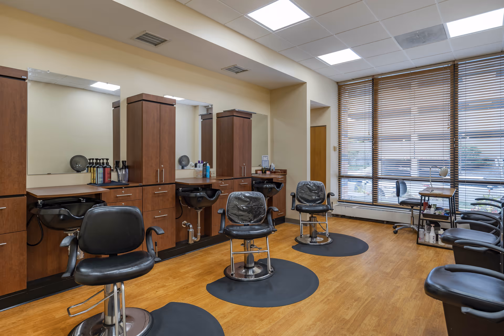 Interior view of a hair salon area with three black salon chairs in front of wooden cabinets and mirrors. The floor is wooden, and there are large windows with wooden blinds letting in natural light. Various hair care products are placed on the counters.